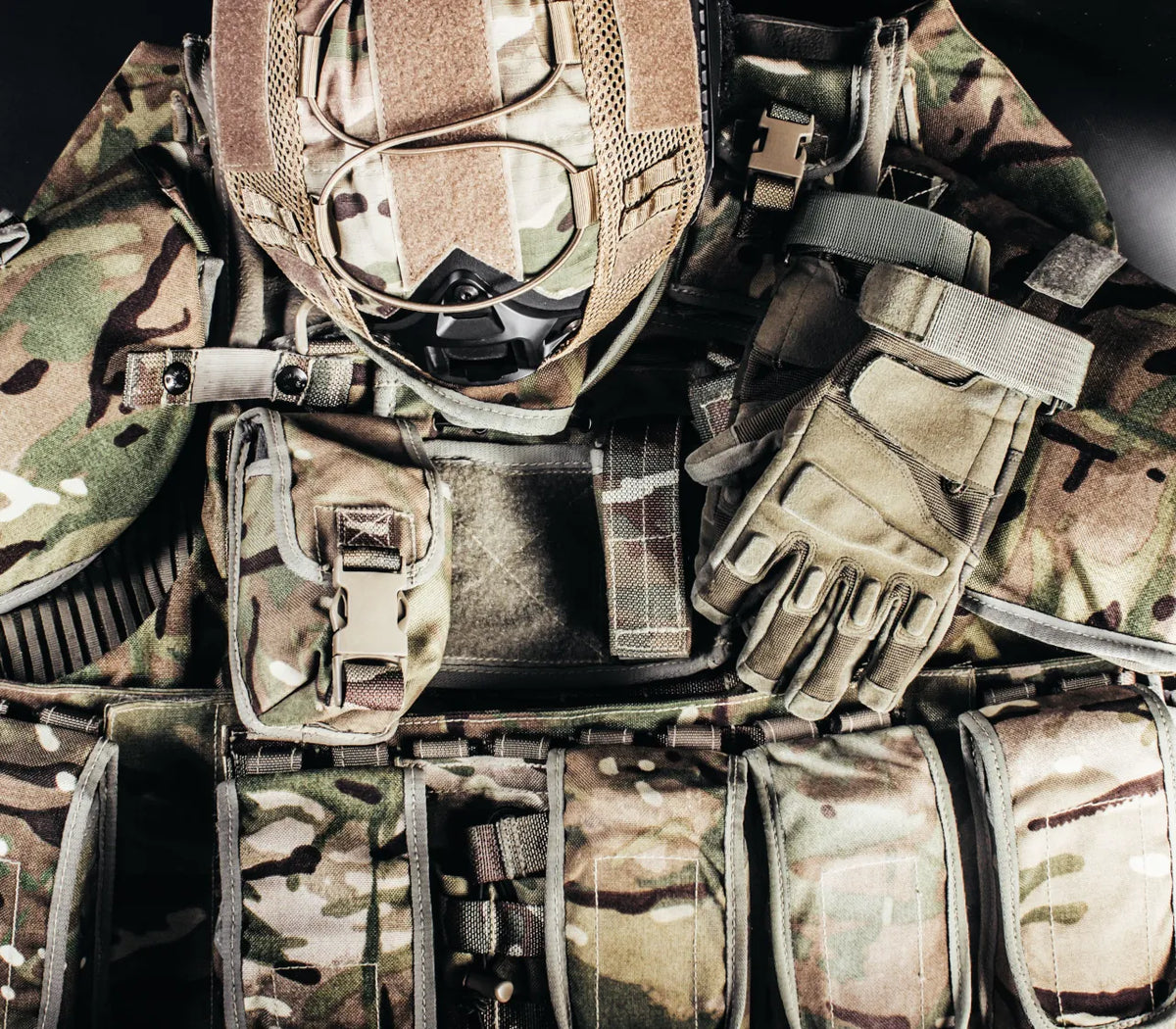 Close-up of tactical gear in camouflage fabric, including a helmet with a face mask, gloves, straps, and utility pouches.